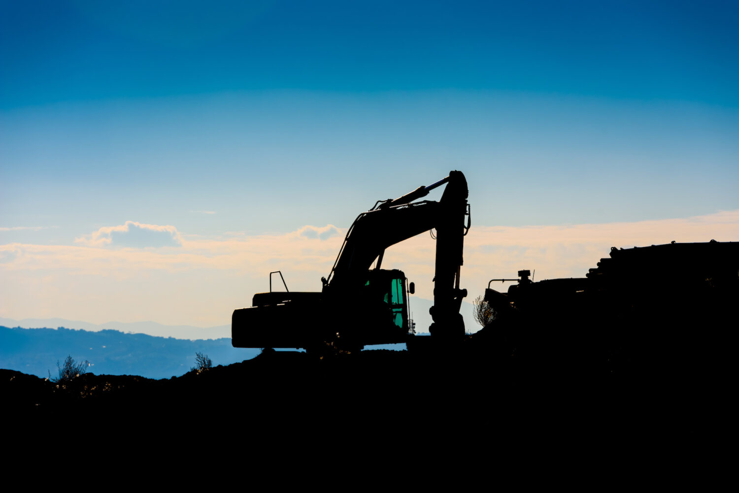 Backlit silhouette of an excavator on top of a hill with unfocused sky background.
